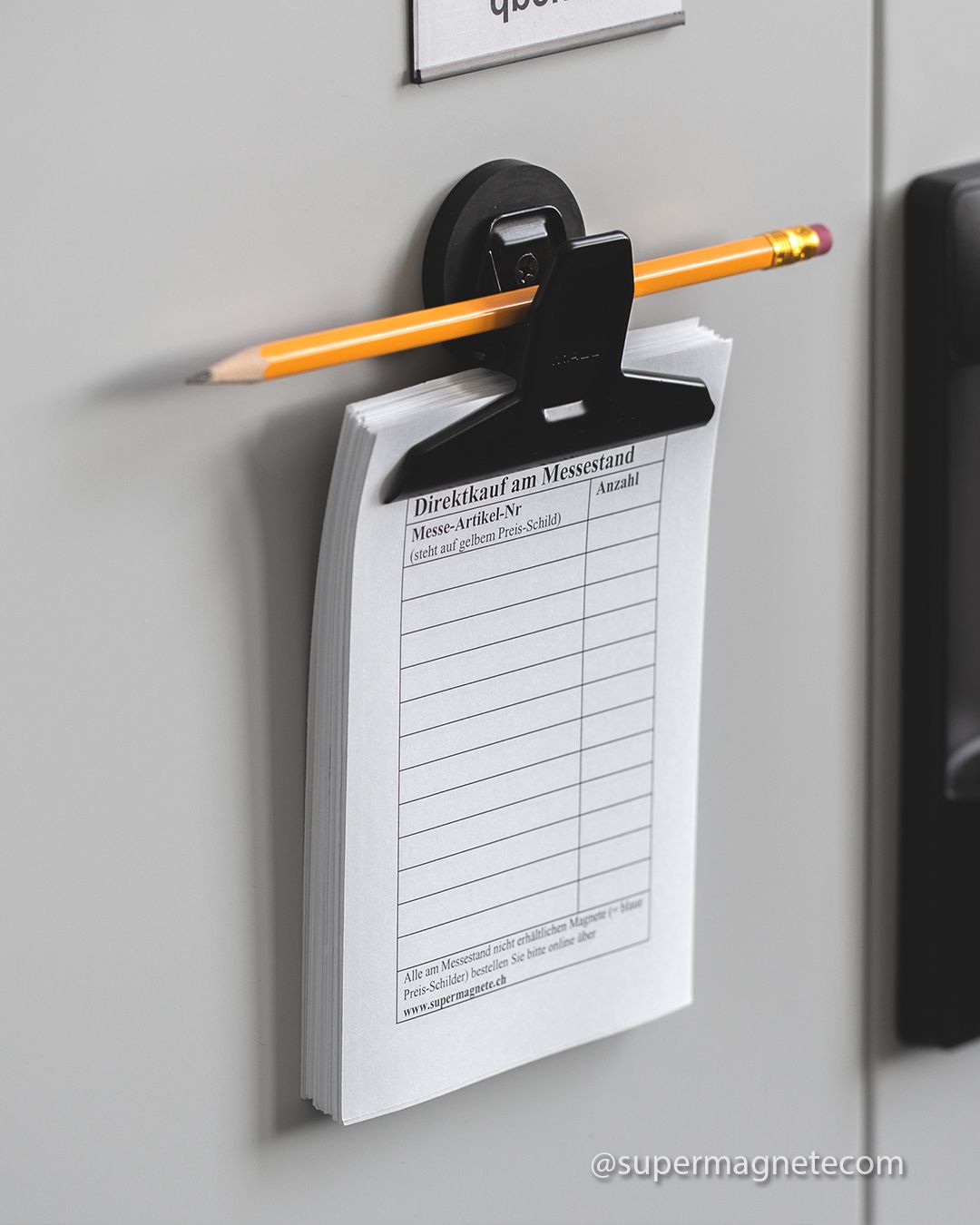 A magnetic clip on a wall holding a printed pad and a pencil